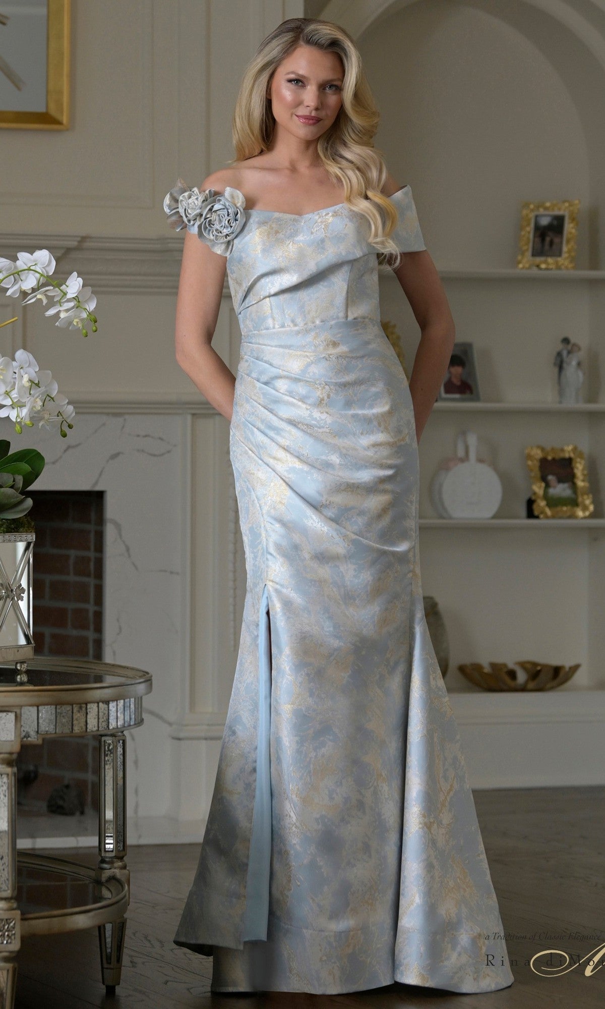 A woman stands indoors in the blue Rina Di Montella RD3522 long formal dress, crafted from jacquard fabric with off-shoulder sleeves and gold floral details; her hands are in pockets, with shelves and a fireplace in the background.